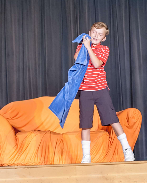 Young boy dressed as Linus from Peanuts on stage acting in front of orange couch